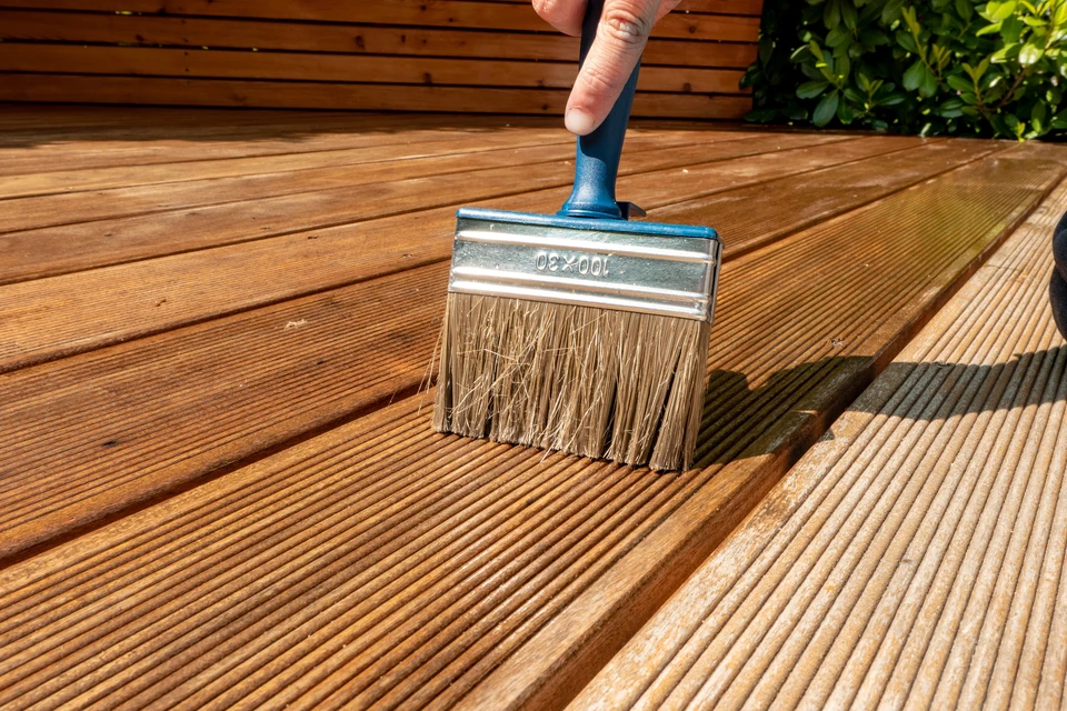 Main ouvrier appliquant un produit de finition sur planches de bois brut avec pinceau plat, portant gants de protection blancs